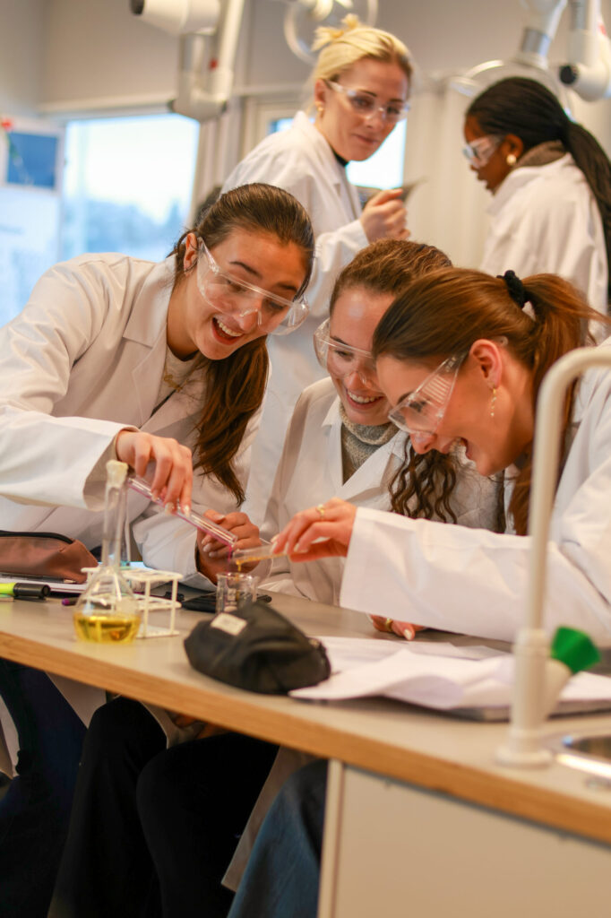 8T1A1715-2 Tre studenter iført laboratoriefrakker og vernebriller utfører et eksperiment sammen ved et laboratoriebord med begerglass og glass, mens andre arbeider i bakgrunnen.