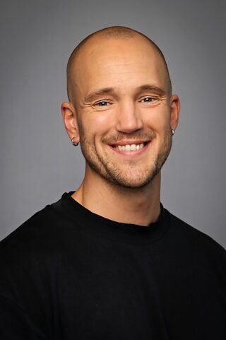 Håkon Øverland Vikse En skallet mann med lys hud og øreringer, iført svart skjorte, smiler til kameraet mot en grå bakgrunn.