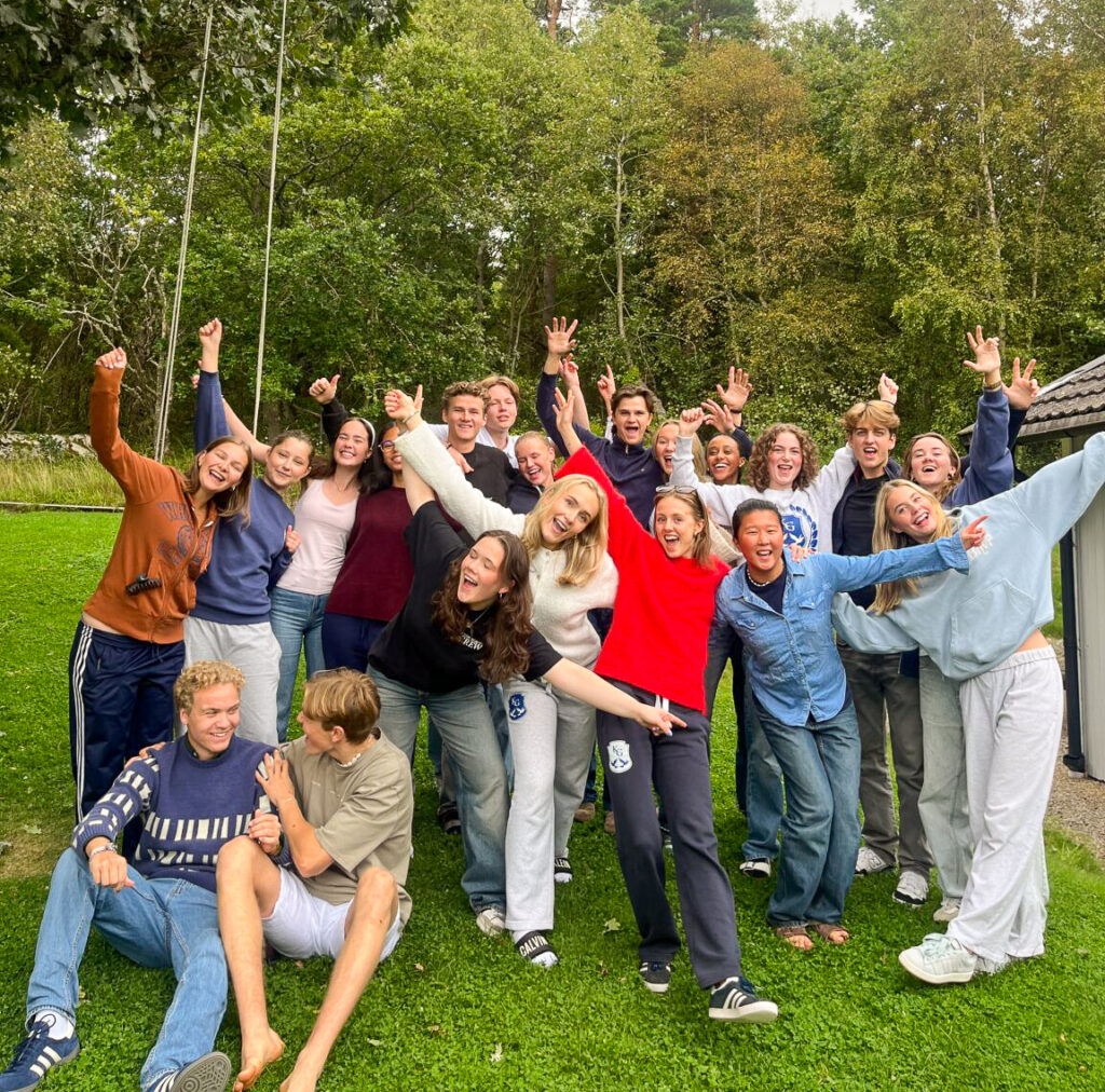 En gruppe mennesker poserer utendørs på gress, med trær i bakgrunnen. Noen sitter og andre står, alle smiler og løfter armene.