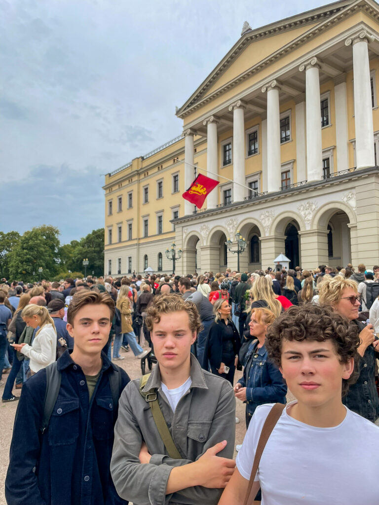 IMG_6743 Tre unge menn står foran en stor folkemengde utenfor en gul nyklassisistisk bygning med søyler og et rødt flagg.