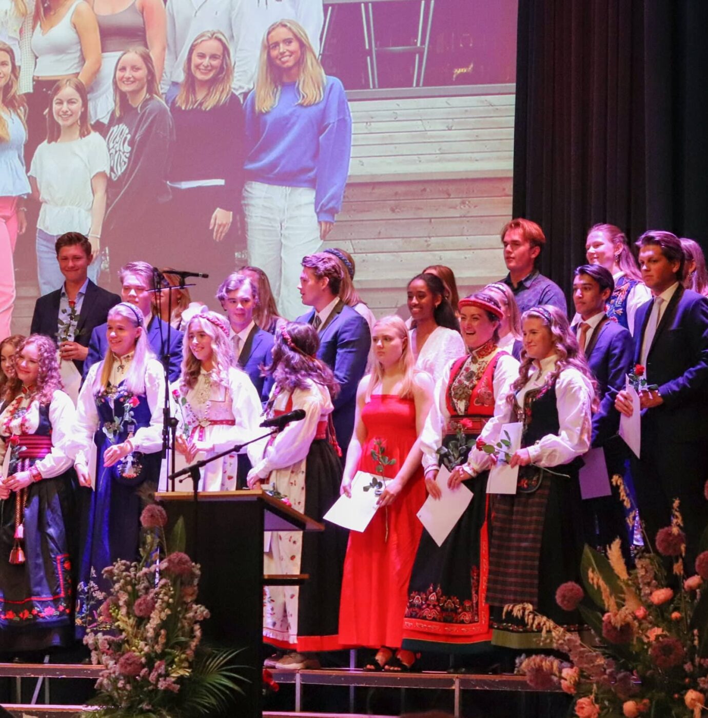 En gruppe unge mennesker kledd i formelle og tradisjonelle klær står på scenen med blomster og papirer, med et projisert bilde bak seg.