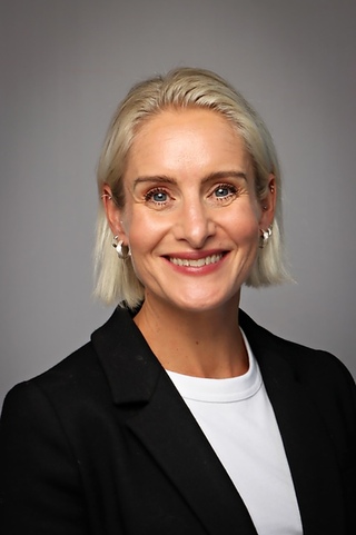 Ragnhild Amlie Svartdahl En kvinne med kort, blondt hår, iført svart blazer og hvit topp, smiler til kameraet mot en grå bakgrunn.