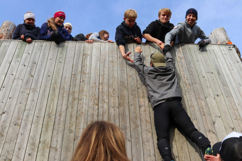 Side 4-5 Vegg-2 En gruppe mennesker hjelper noen med å klatre over en trevegg og strekker seg ned for å gi en hånd.