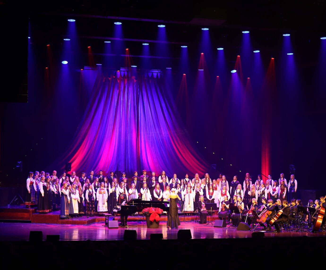 Et kor og et orkester opptrer på scenen under lilla og rød belysning, med en pianist og en solosanger i forgrunnen.