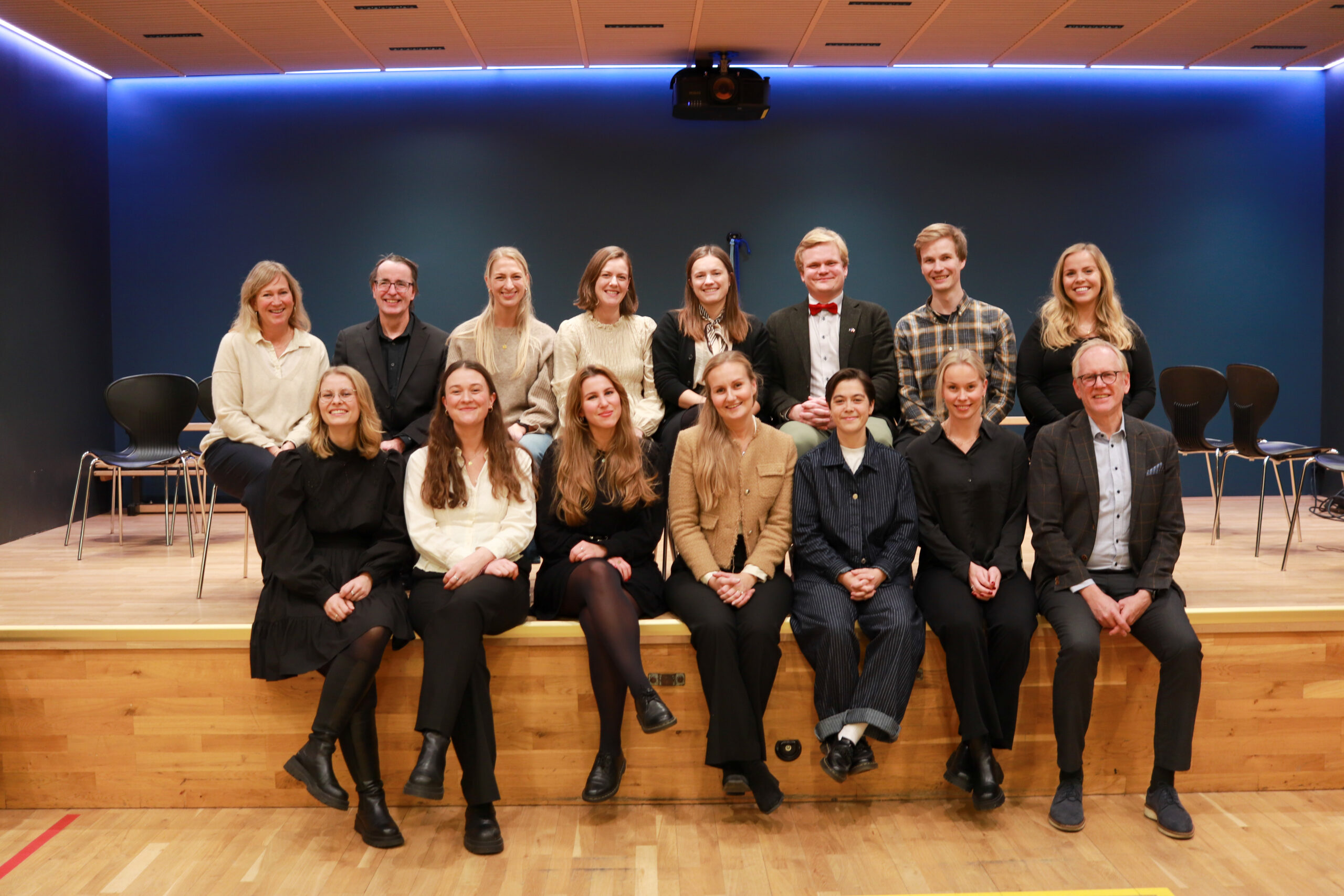 En gruppe på femten personer, kledd i halvformelle antrekk, sitter og står på en scene foran en tom stolrekke og en blå vegg.