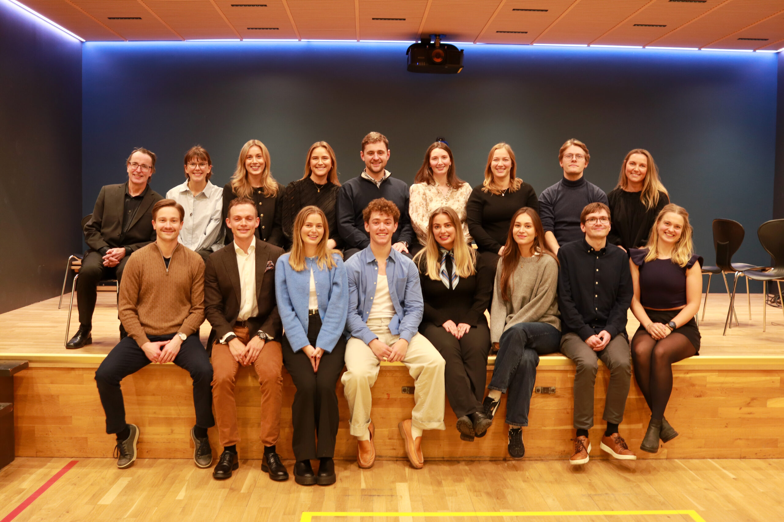 Sytten personer poserer for et gruppebilde i to rader på en trescene i et rom med blå vegger og taklamper.