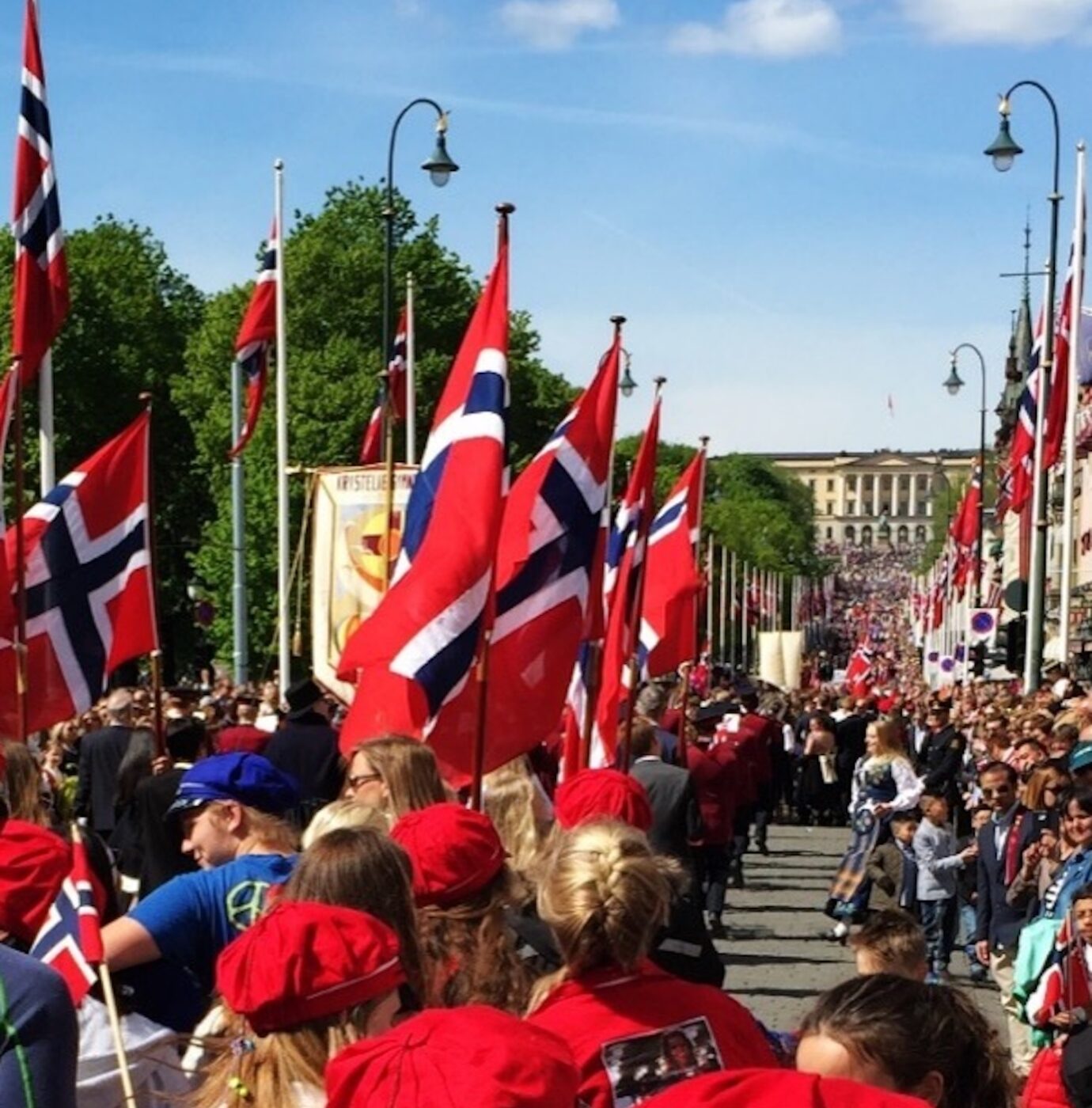 En stor folkemengde, mange med røde luer, går nedover en gate kantet med norske flagg under en parade på en solskinnsdag.