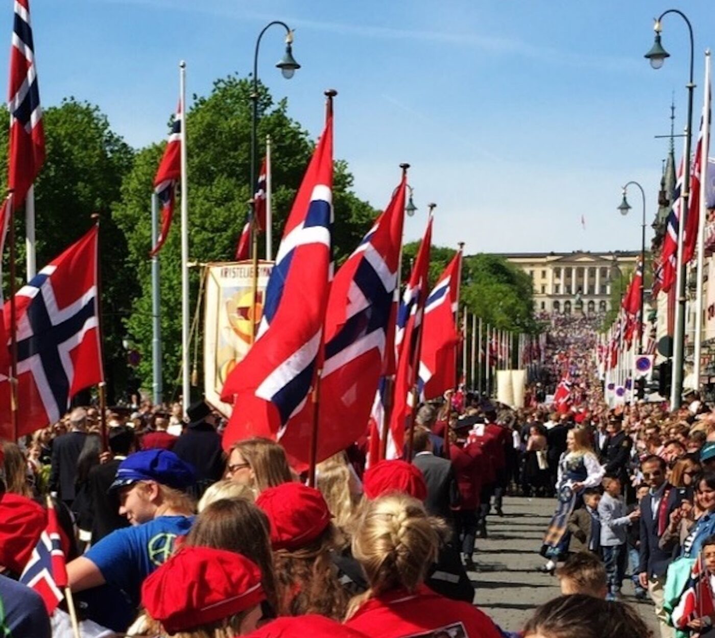 En stor folkemengde, mange med røde luer, går nedover en gate kantet med norske flagg under en parade på en solskinnsdag.