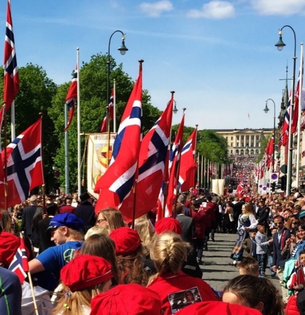 17mai_forside En stor folkemengde, mange med røde luer, går nedover en gate kantet med norske flagg under en parade på en solskinnsdag.