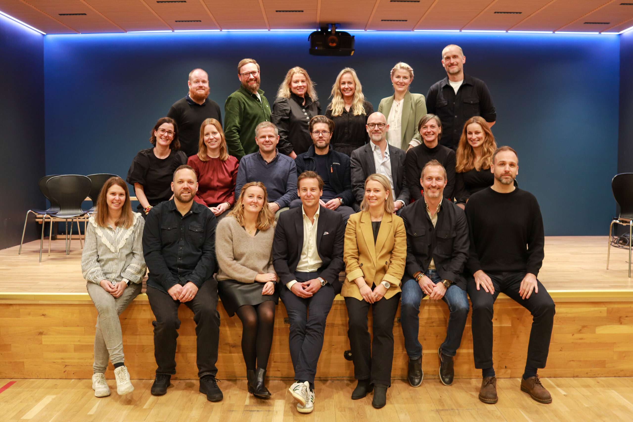 En gruppe på 22 personer poserer for et bilde på en trescene i tre rader, med en blå vegg og tomme stoler i bakgrunnen.