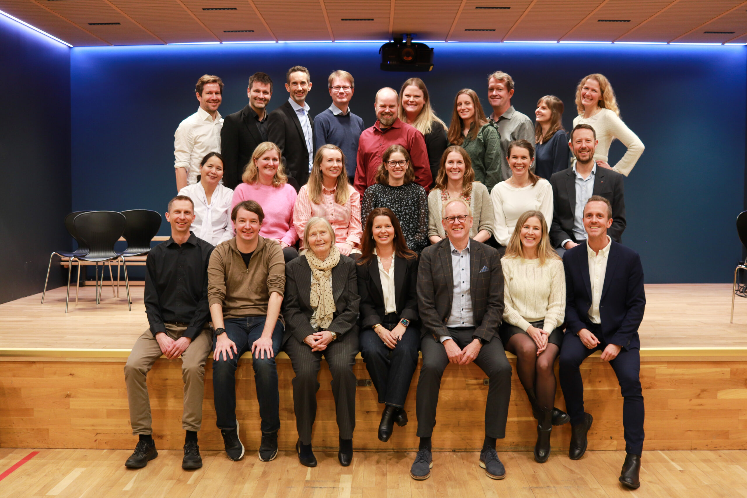 En gruppe på 22 personer, oppstilt i tre rader, poserer og smiler for et gruppebilde på en scene med tregulv og blå bakgrunn.