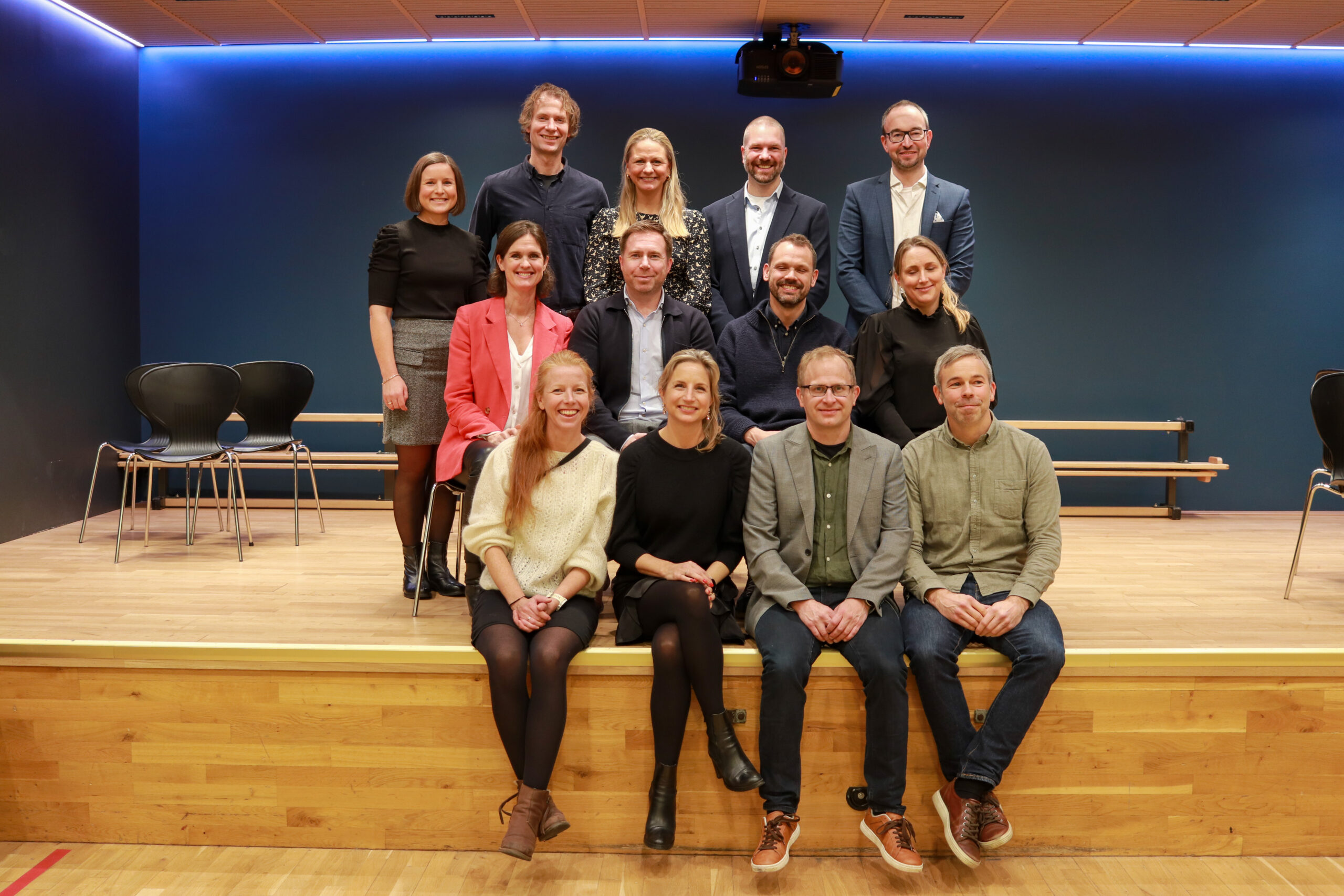 Fjorten personer poserer for et gruppebilde på en scene med tregulv og blå bakgrunn. Noen sitter foran, mens andre står bak dem.