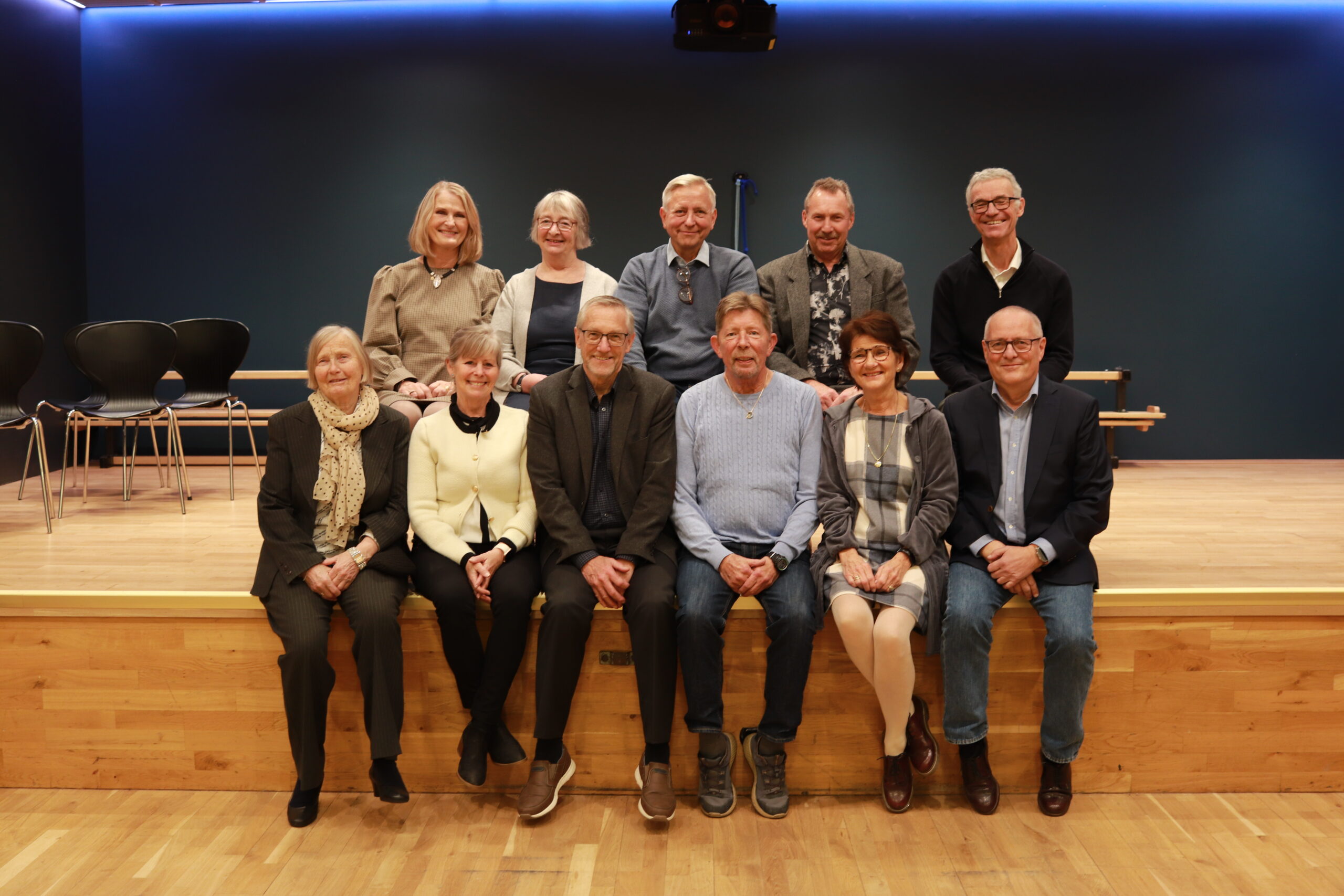 Elleve voksne poserer for et gruppebilde på en trescene, med noen sittende foran og andre stående bak, mot en mørk vegg.