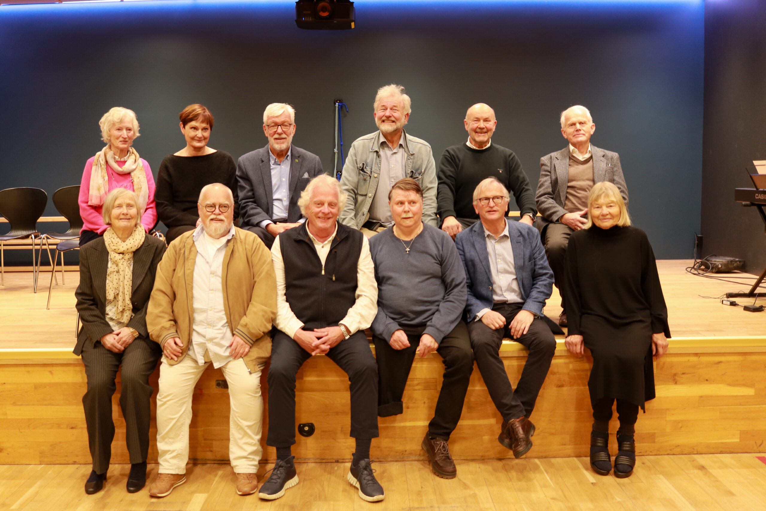 Tretten eldre voksne, både menn og kvinner, sitter og står på en trescene foran en blå vegg og poserer for et gruppebilde.