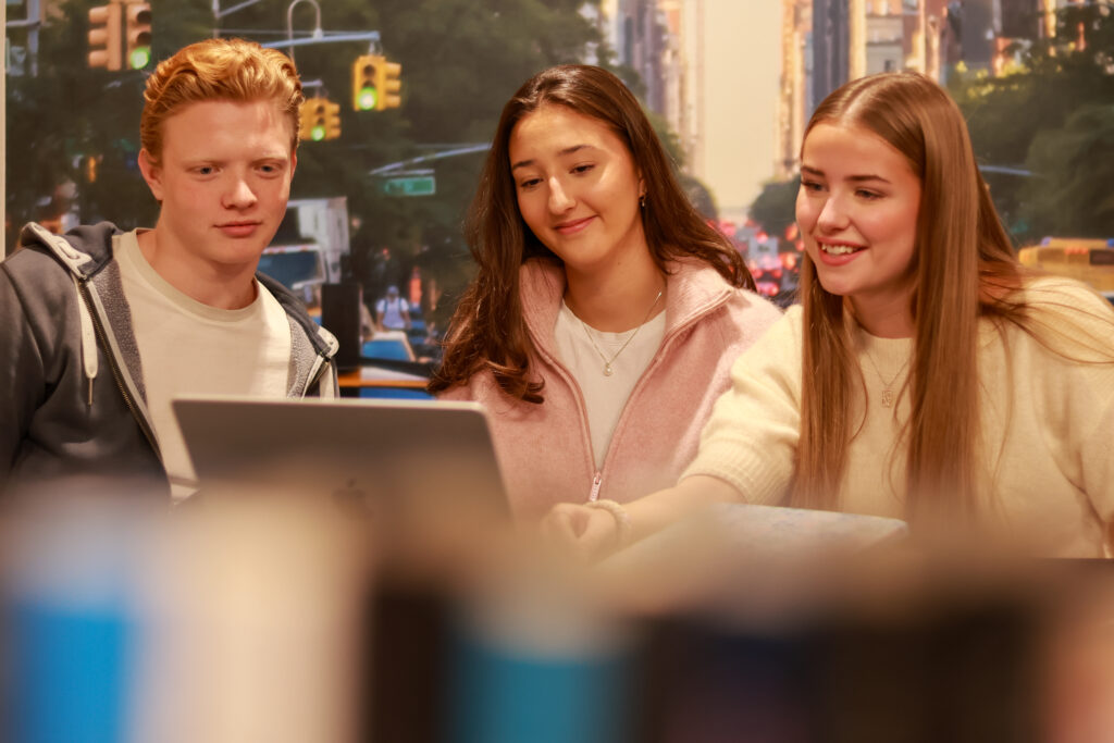 8T1A0064-2 Tre unge voksne sitter sammen og ser på en bærbar datamaskin, de virker fokuserte og engasjerte, med et gatebilde fra en by i bakgrunnen.