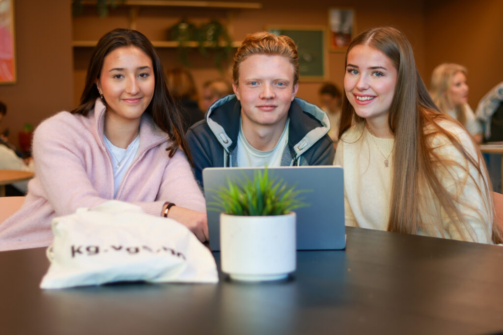 8T1A0190-2 Tre unge voksne sitter sammen ved et bord med en bærbar datamaskin og en potteplante og ser inn i kameraet i et klasserom eller en studiesituasjon.