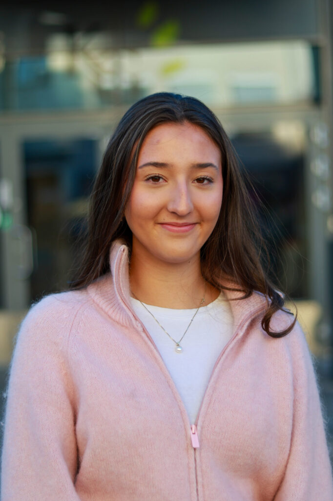 8T1A0194 En ung kvinne med langt brunt hår, iført en lyserosa genser med glidelås og hvit topp, står utenfor, ser inn i kameraet og smiler lett.