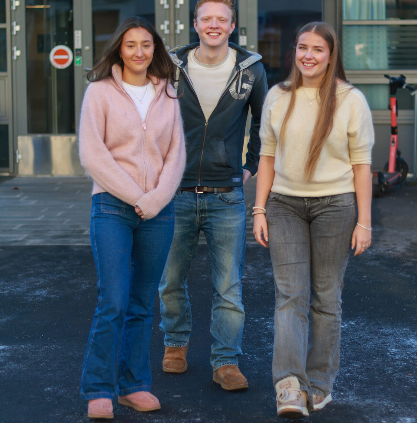 Tre unge voksne, to kvinner og en mann, står utenfor en moderne bygning på et asfaltert område og smiler til kameraet.