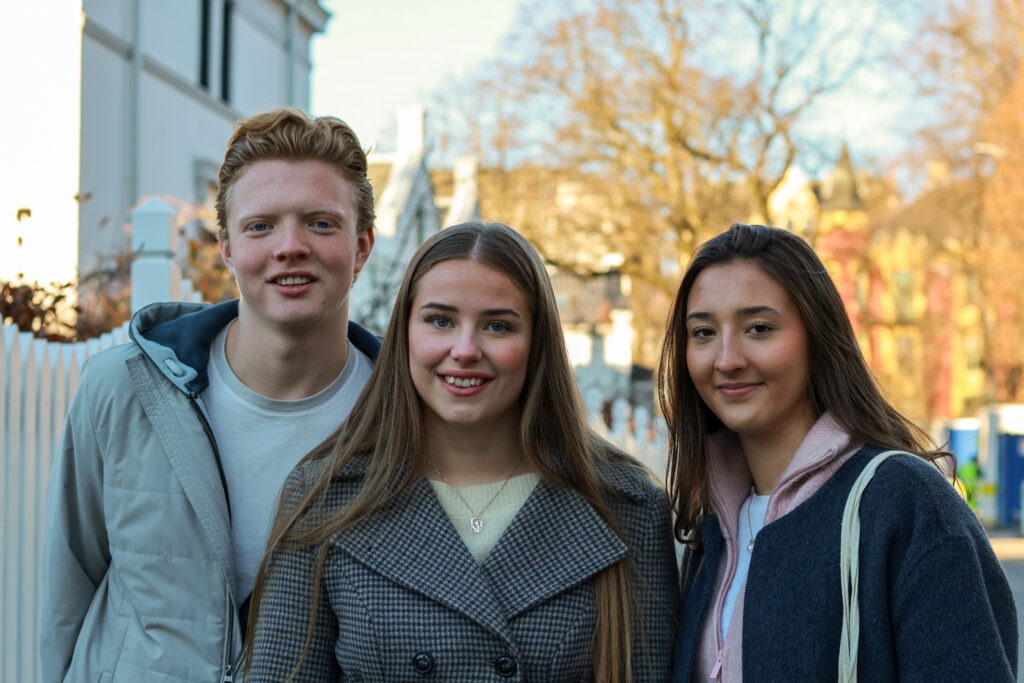 8T1A0304-2 Tre unge voksne står ute en solskinnsdag og smiler til kameraet, med trær og bygninger synlige i bakgrunnen.