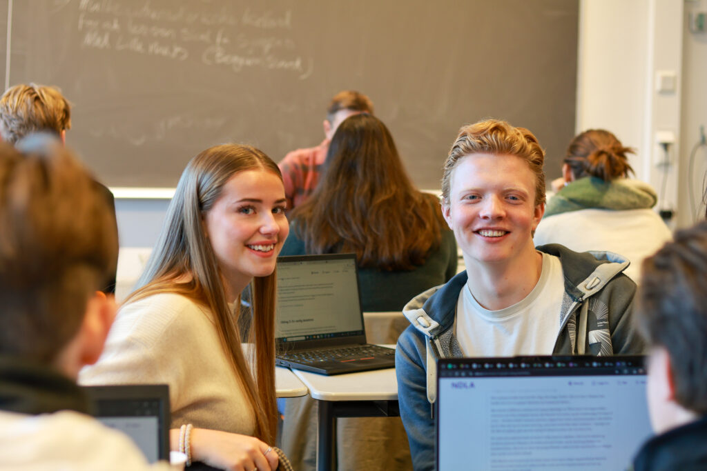8T1A0403-2 To studenter sitter ved pulter i et klasserom og smiler til kameraet, med laptoper foran seg og en tavle i bakgrunnen.