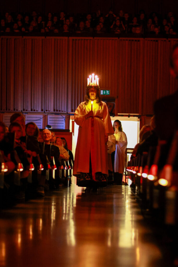 8T1A1015-2 En person iført hvit kjortel og en krone av stearinlys går ned en svakt opplyst midtgang, omgitt av sittende mennesker i et auditorium.