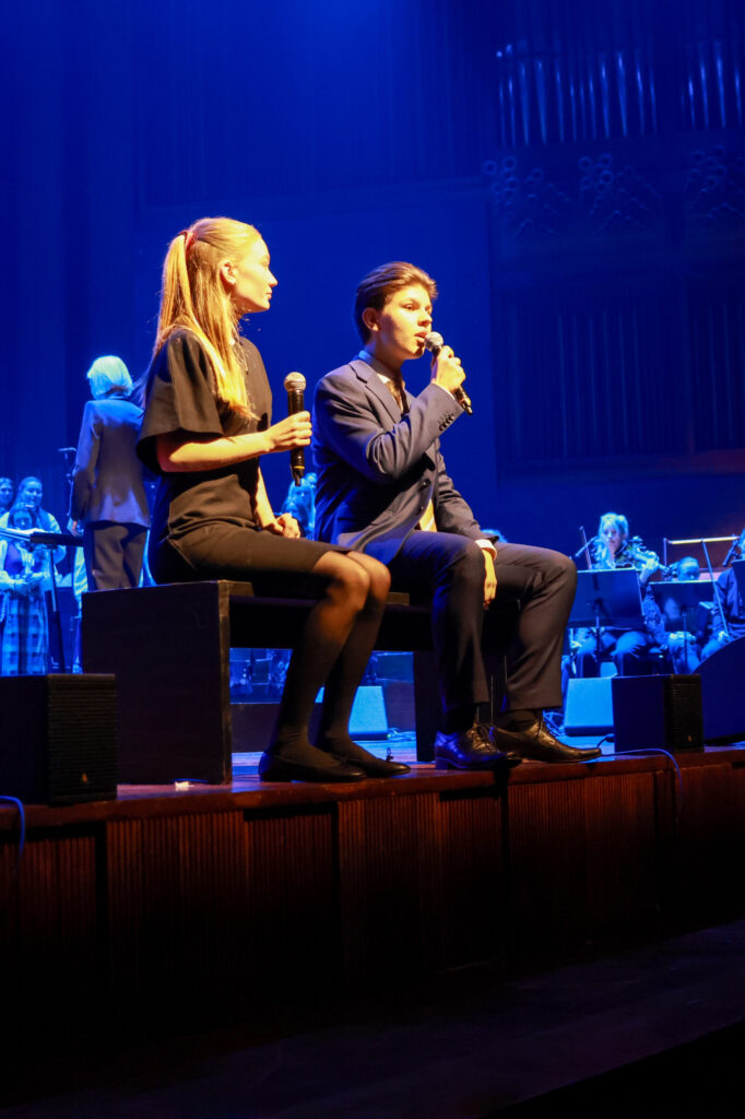 8T1A1195-3 To personer sitter på scenen med mikrofoner i hendene, mens et orkester spiller i bakgrunnen under blå scenelys.