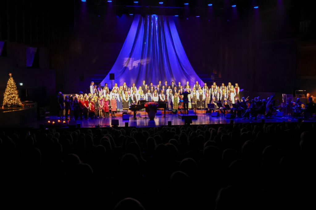 8T1A1349-2 Et kor opptrer på en scene med blå belysning og draperte stoffer i bakgrunnen; et pyntet juletre er synlig ved siden av, og musikerne sitter til høyre.