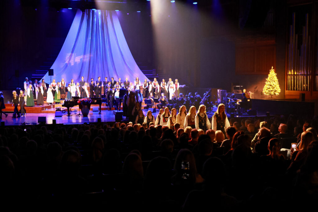 8T1A1351-2 En sceneforestilling med et stort kor og orkester, dramatisk lyssetting, et opplyst juletre og en gruppe mennesker som går ned midtgangen mot scenen.