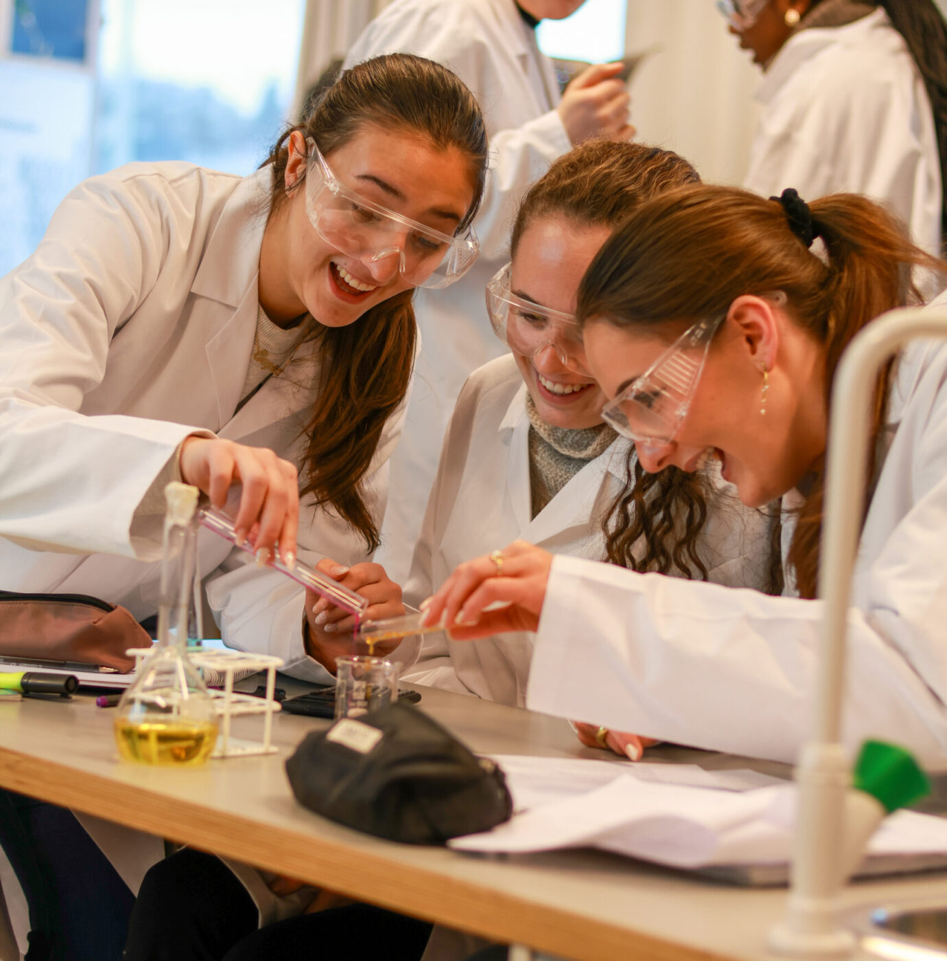 8T1A1715-2 Tre studenter iført laboratoriefrakker og vernebriller utfører et eksperiment sammen ved et laboratoriebord med begerglass og glass, mens andre arbeider i bakgrunnen.