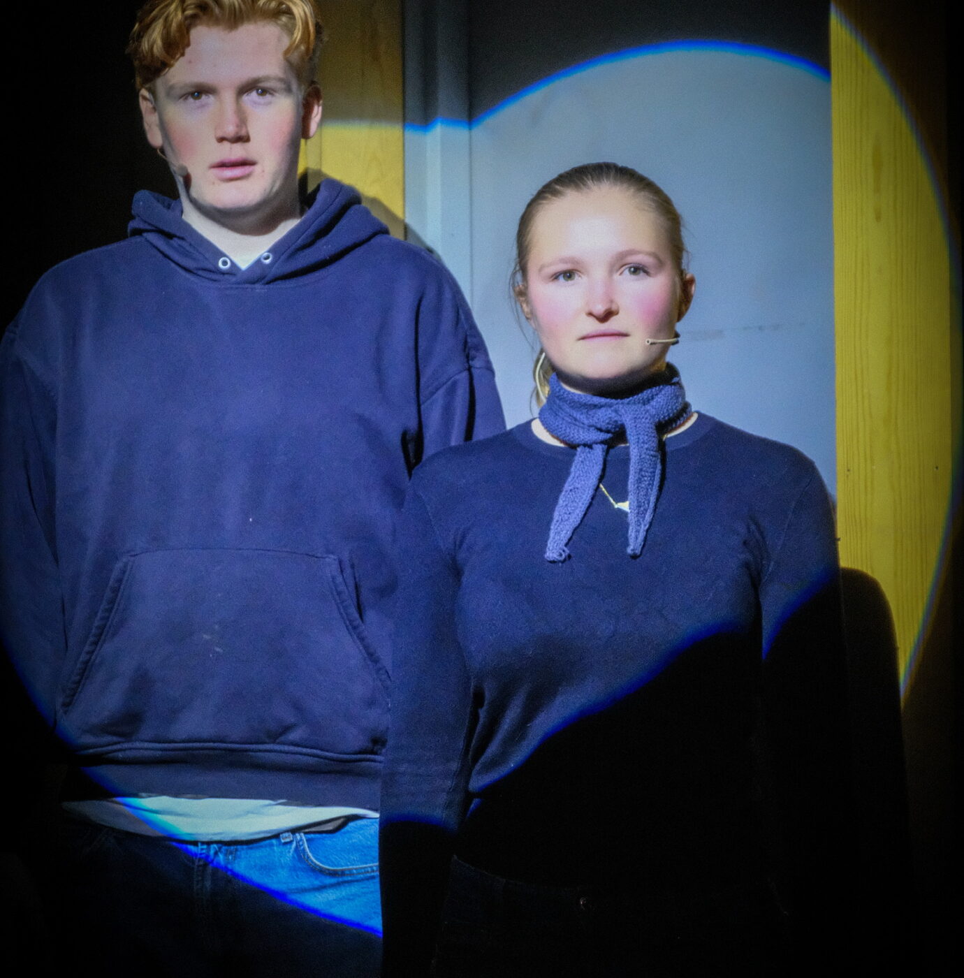 DSCF0841 To unge mennesker, en mann og en kvinne, står side om side foran en lukket dør, opplyst av en spotlight i en ellers mørk omgivelse.