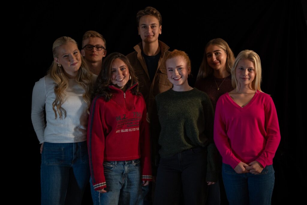 En gruppe på seks unge mennesker som står sammen på rekke foran en svart bakgrunn.