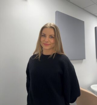 En kvinne med langt blondt hår og svart genser står innendørs foran grå akustiske paneler, vendt mot kameraet og smiler lett.