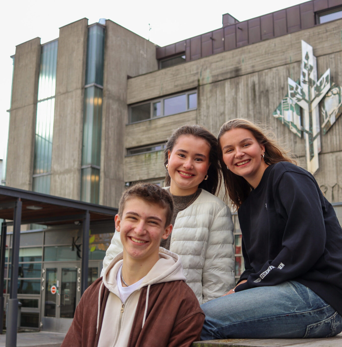 IMG_2400-2 Tre personer smilende og sittende utenfor en betongbygning med moderne design og treskulptur på fasaden.