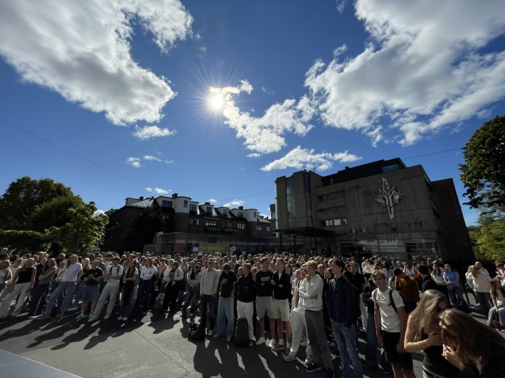 En stor gruppe mennesker står utendørs under en delvis overskyet himmel, med sollys som skinner ned, i nærheten av moderne bygninger og trær.