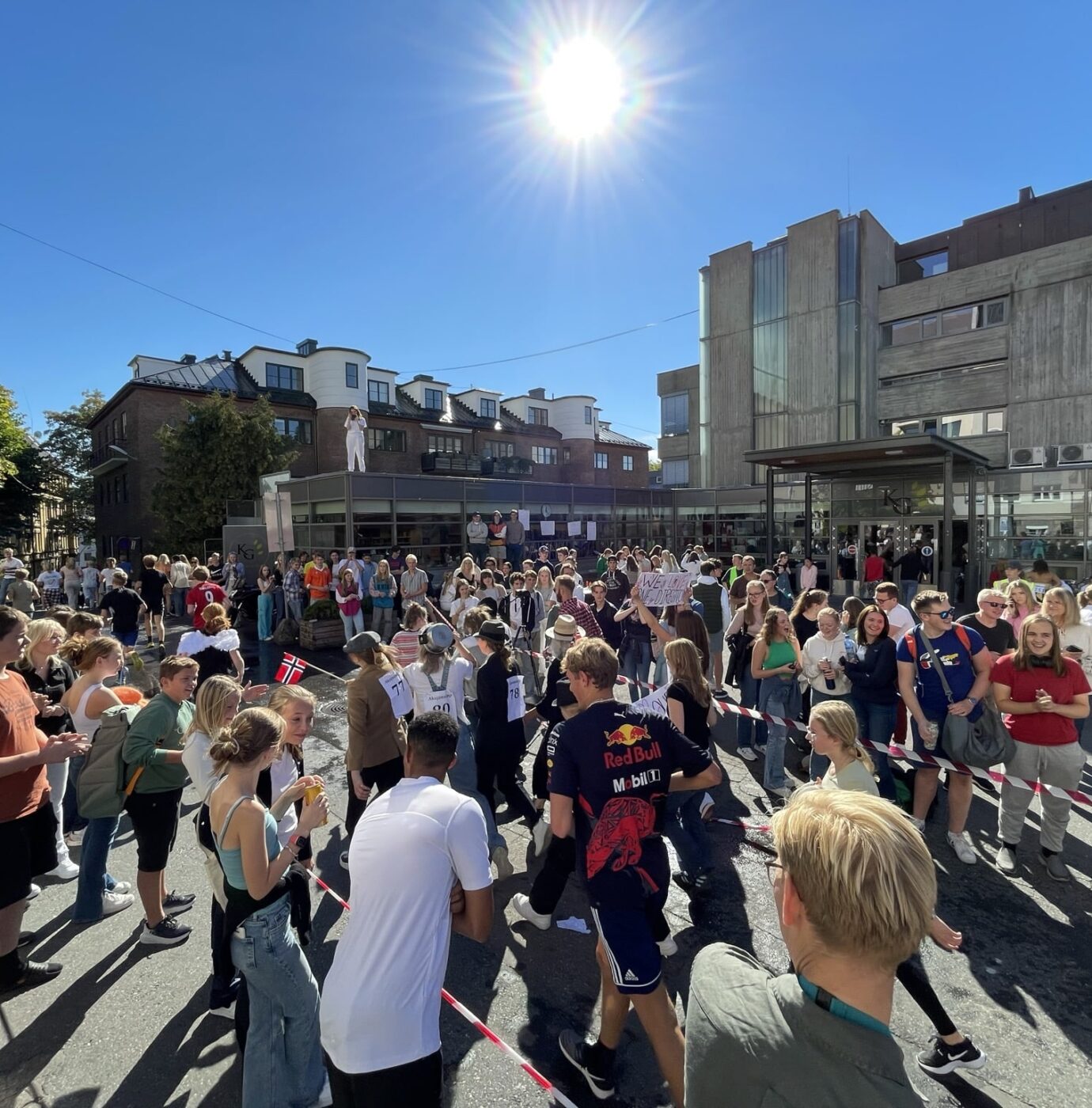 En stor gruppe mennesker samles utendørs i et solfylt byområde, noen holder flagg og ballonger. Bygninger og trær er i bakgrunnen under en klar blå himmel.
