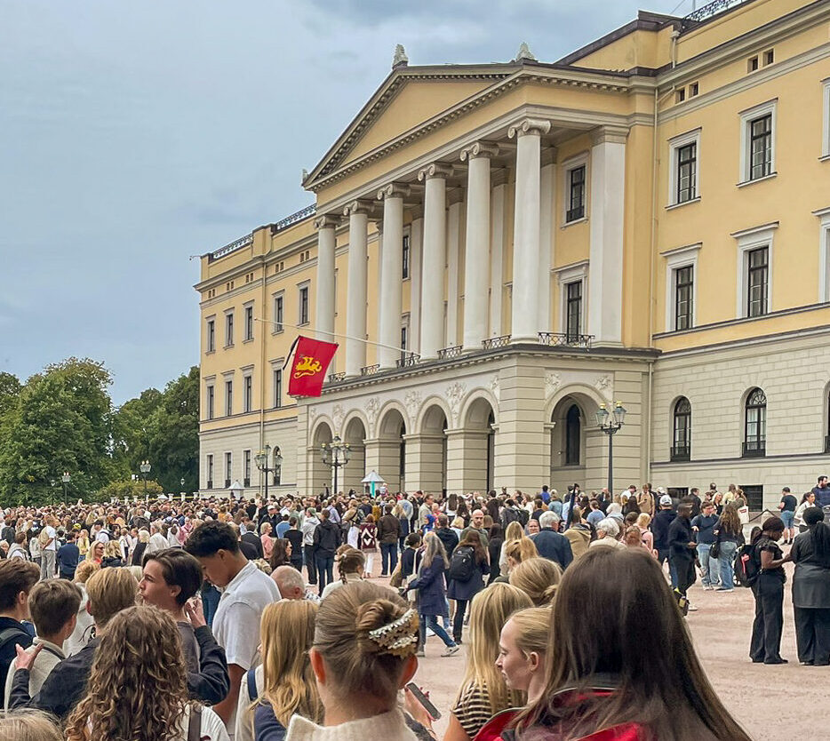 IMG_6736-2 En stor folkemengde samler seg utenfor en gul nyklassisistisk bygning med søyler og en balkong, med et rødt og gullfarget flagg hengt opp på fasaden.