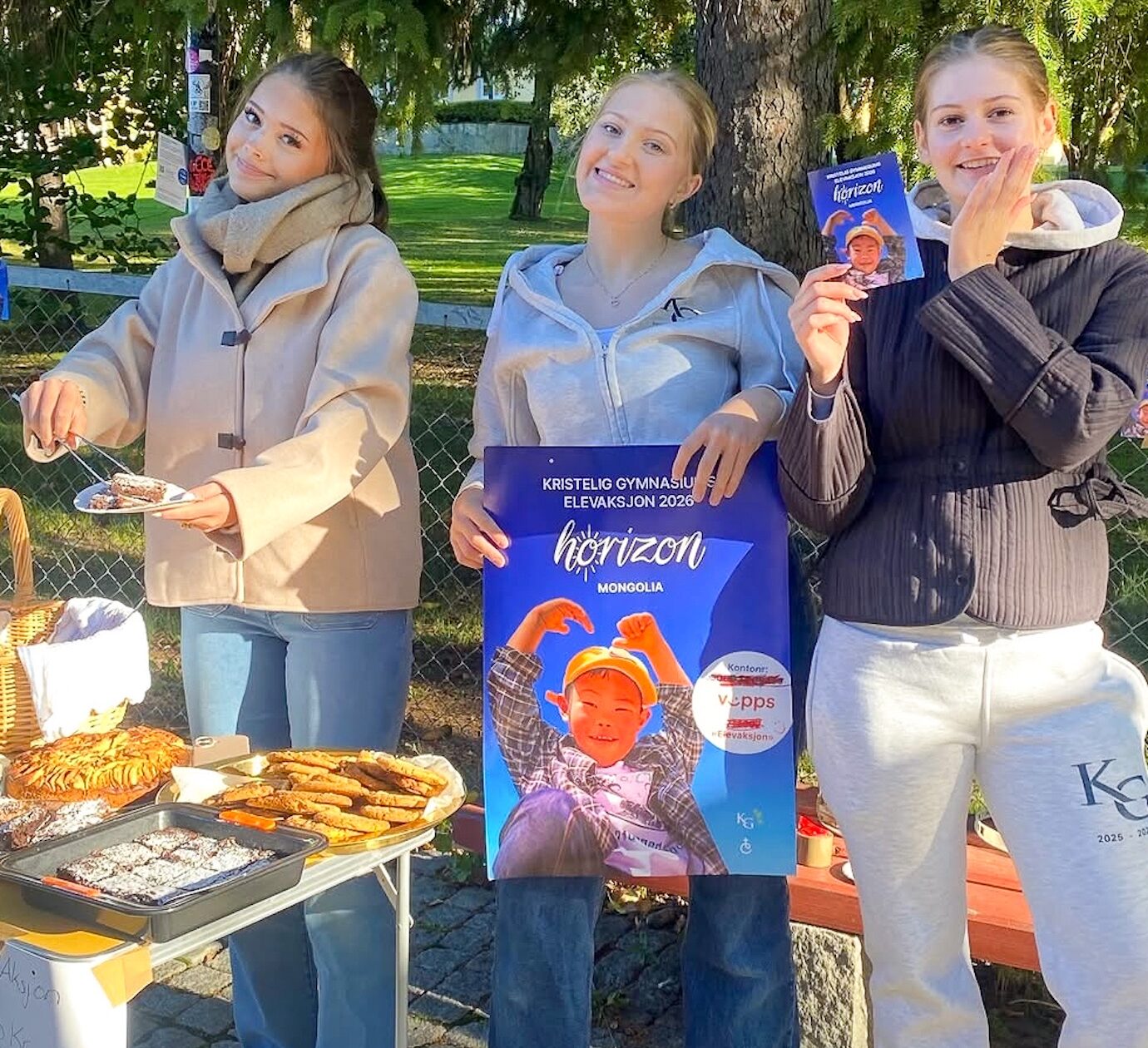 Tre unge kvinner står bak et bord med bakverk og holder opp og viser frem en stor plakat og et flygeblad for et arrangement kalt «horizon» utendørs på en solrik dag.