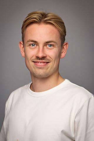 Ole Martin Flaat En ung mann med kort blondt hår og lys bart, iført hvit T-skjorte, smiler til kameraet mot en grå bakgrunn.