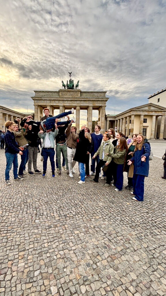 Side 4-5 Berlin 2 En gruppe mennesker poserer foran Brandenburger Tor; flere løfter en person horisontalt, mens andre smiler og gestikulerer mot kameraet.