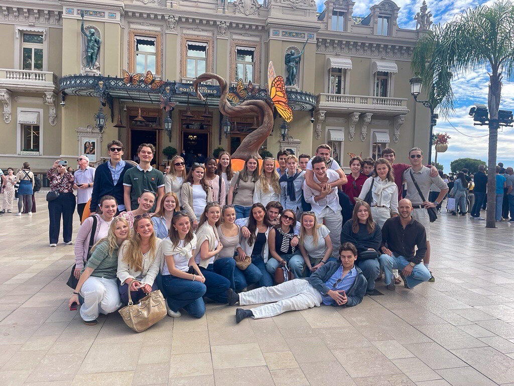 Side 4-5 Nice 2 En stor gruppe unge voksne poserer foran en utsmykket bygning med en treskulptur og sommerfugldekorasjoner.