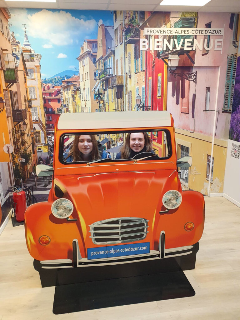 Side 4-5 Nice 3 To kvinner poserer bak et utsnitt av en oransje bil foran en Provence-kulisse med fargerike bygninger og et