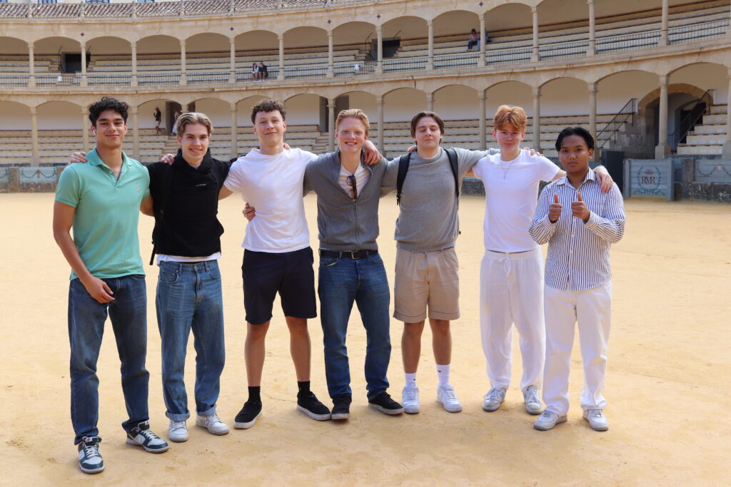 Side 5-7 Malaga 2 Sju unge menn står på rekke og rad på en sandflate ved en tom tyrefekterarena og poserer for et gruppebilde med armene rundt hverandre.