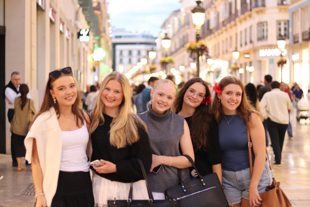 Side 5-7Malaga 1 Fem unge kvinner står tett sammen og smiler når de skal fotograferes i en travel, godt opplyst gågate med butikker og mennesker i bakgrunnen.