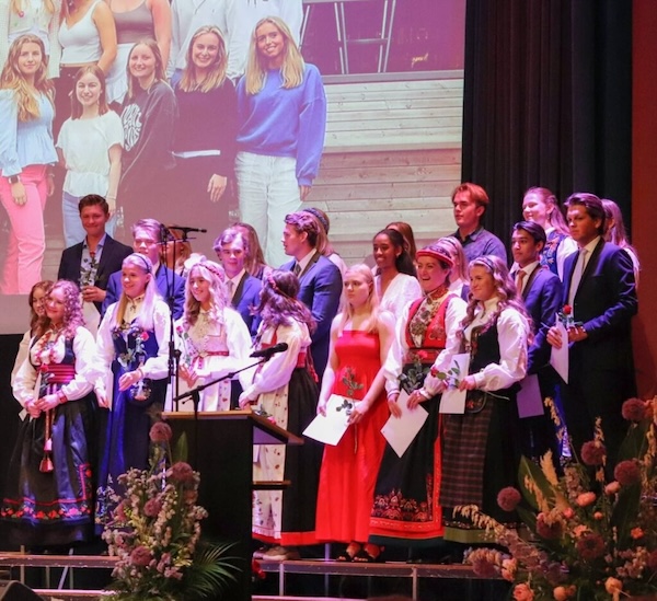 avslutning_lite En gruppe unge voksne i formelle og tradisjonelle antrekk står på scenen med blomster og diplomer i hendene, med et projisert gruppebilde i bakgrunnen.